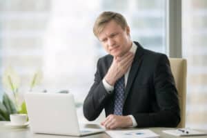 Young businessman at his desk holding his neck in discomfort while working on a laptop, showing symptoms of sore throat, voice loss, and irritation from vocal strain or illness.