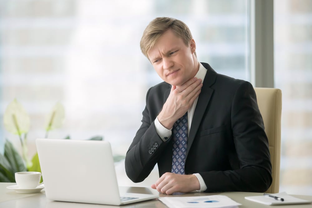 Young businessman at his desk holding his neck in discomfort while working on a laptop, showing symptoms of sore throat, voice loss, and irritation from vocal strain or illness.